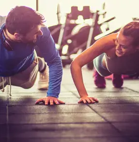 A man and a woman doing push-ups together in a gym, both smiling and engaging in a friendly competition.