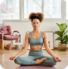 A woman in a serene yoga pose, sitting cross-legged on a mat in a well-lit room with plants and large windows.